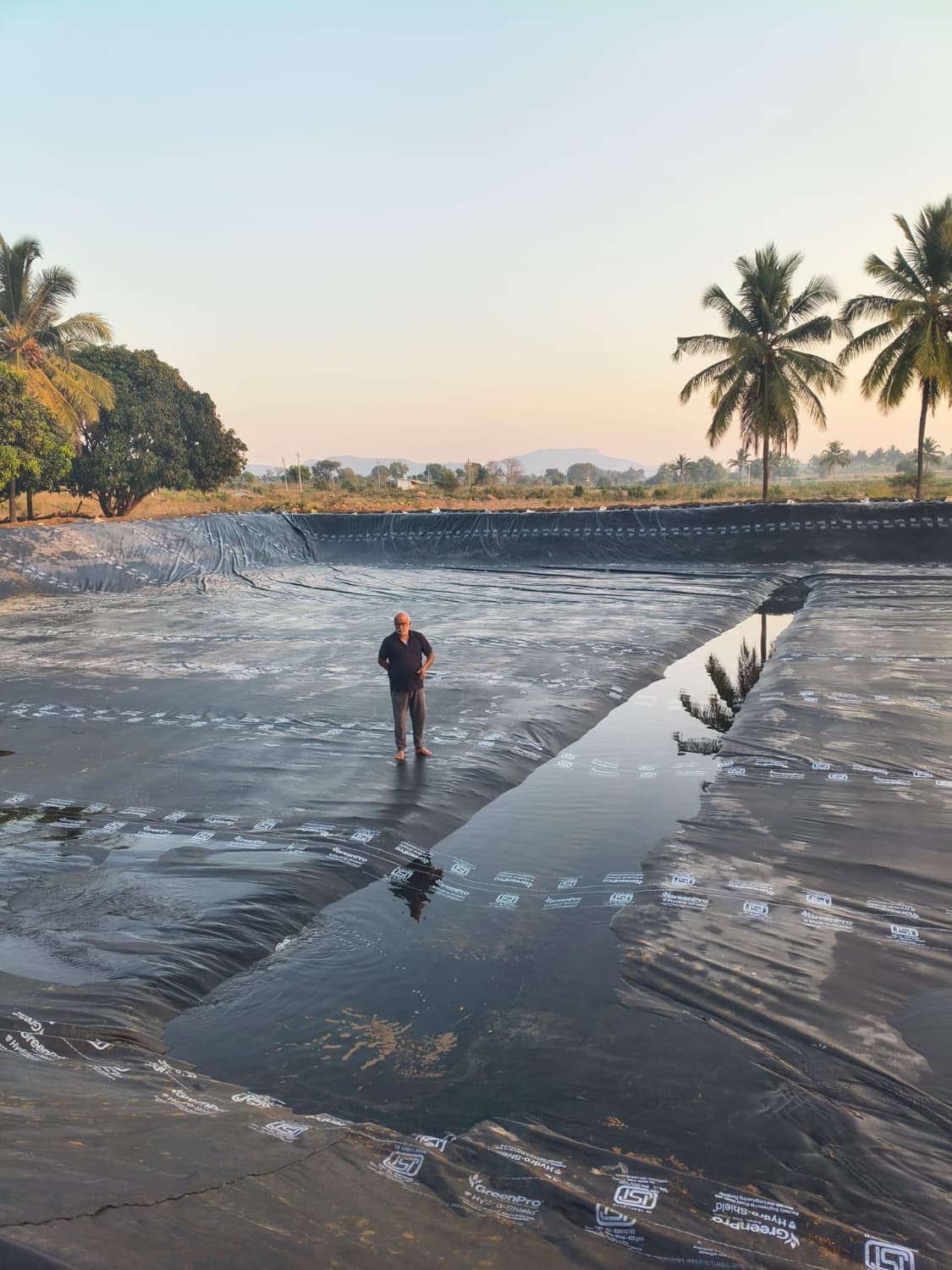 A farmer stands on the fresh black liner as the first water begins to pool in the new pond at dusk