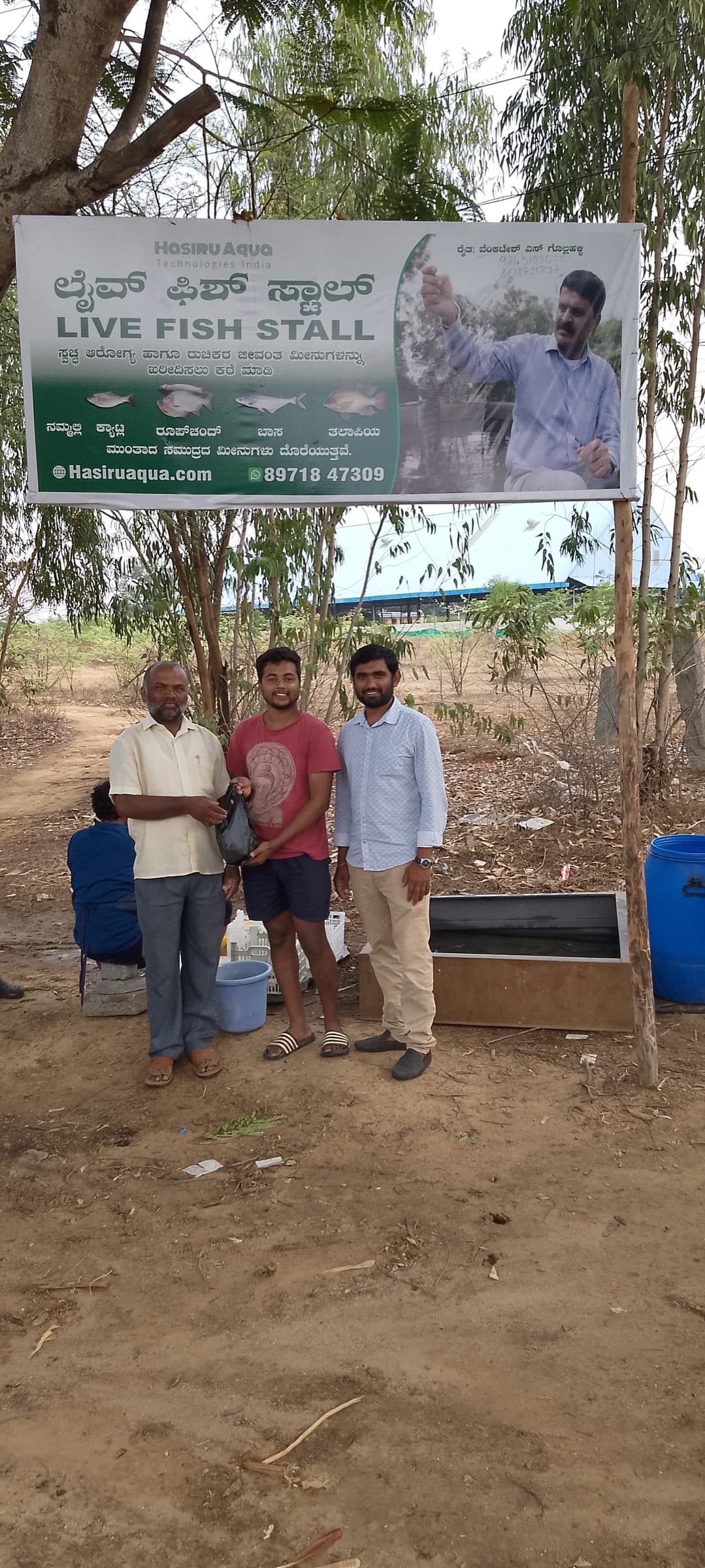 Three men stand under a HasiruAqua LIVE FISH STALL banner at a farm-side live-fish stand