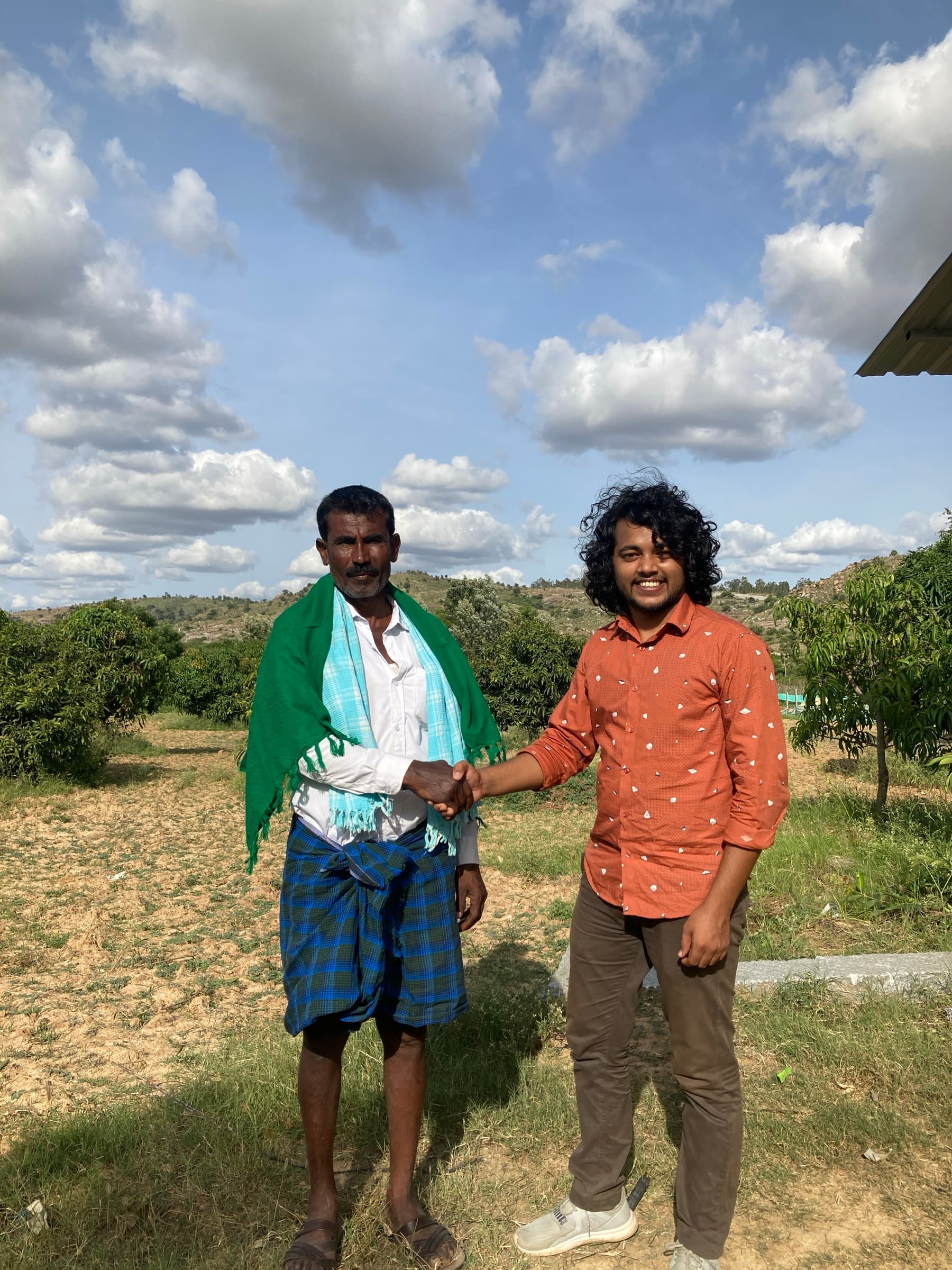 A HasiruAqua founder and a farmer in a green shawl shake hands at the edge of a field under a big sky